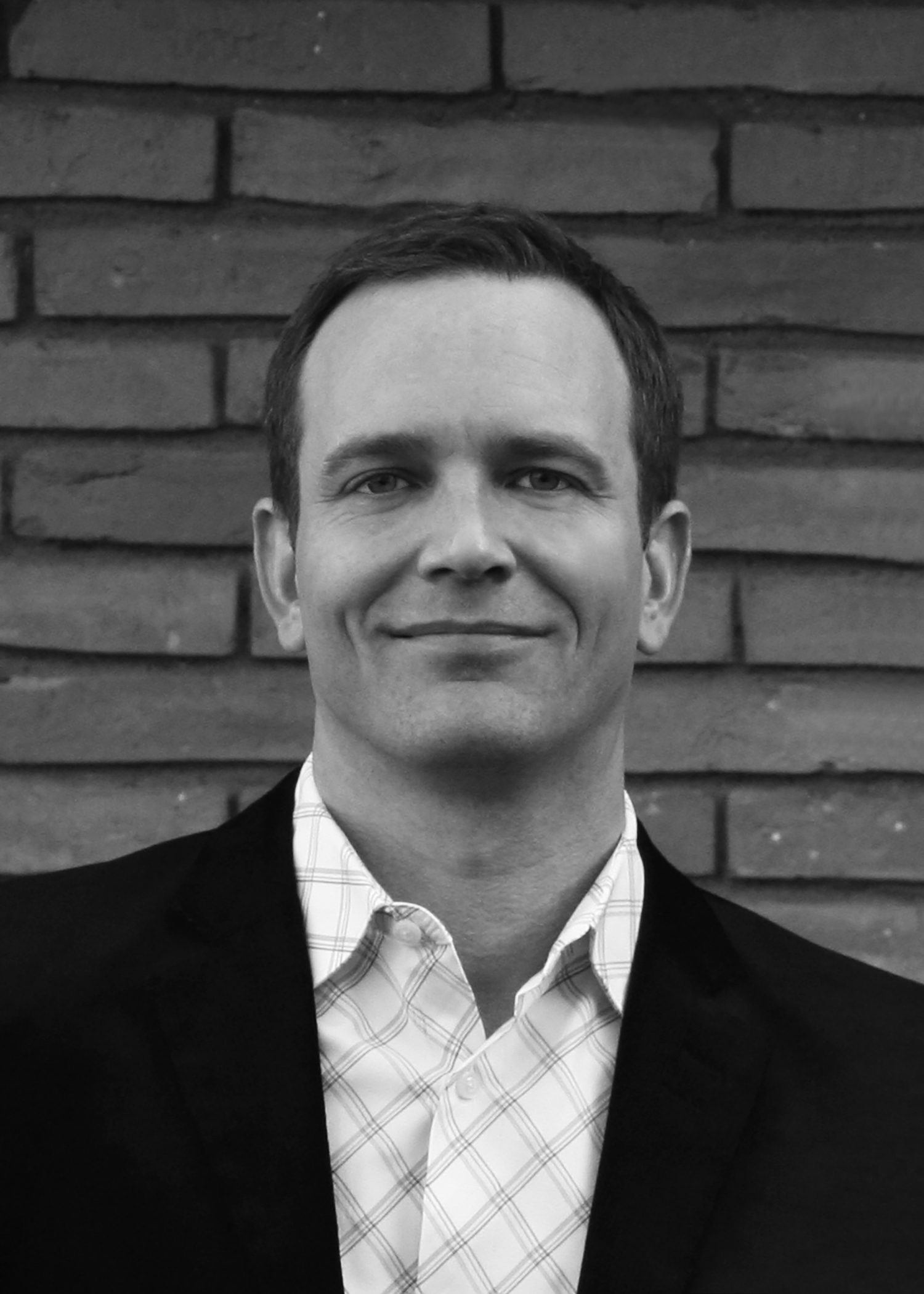 A man in a black jacket over a checked shirt smiles slightly in front of a brick wall.