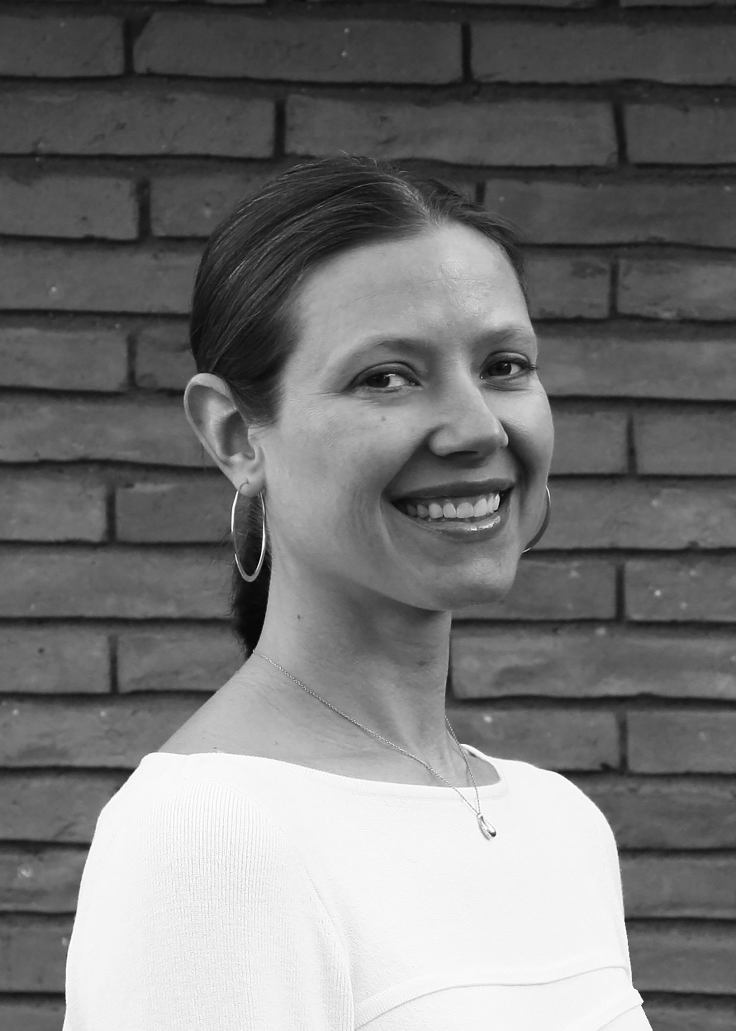 A smiling woman in a white top standing against a brick wall.