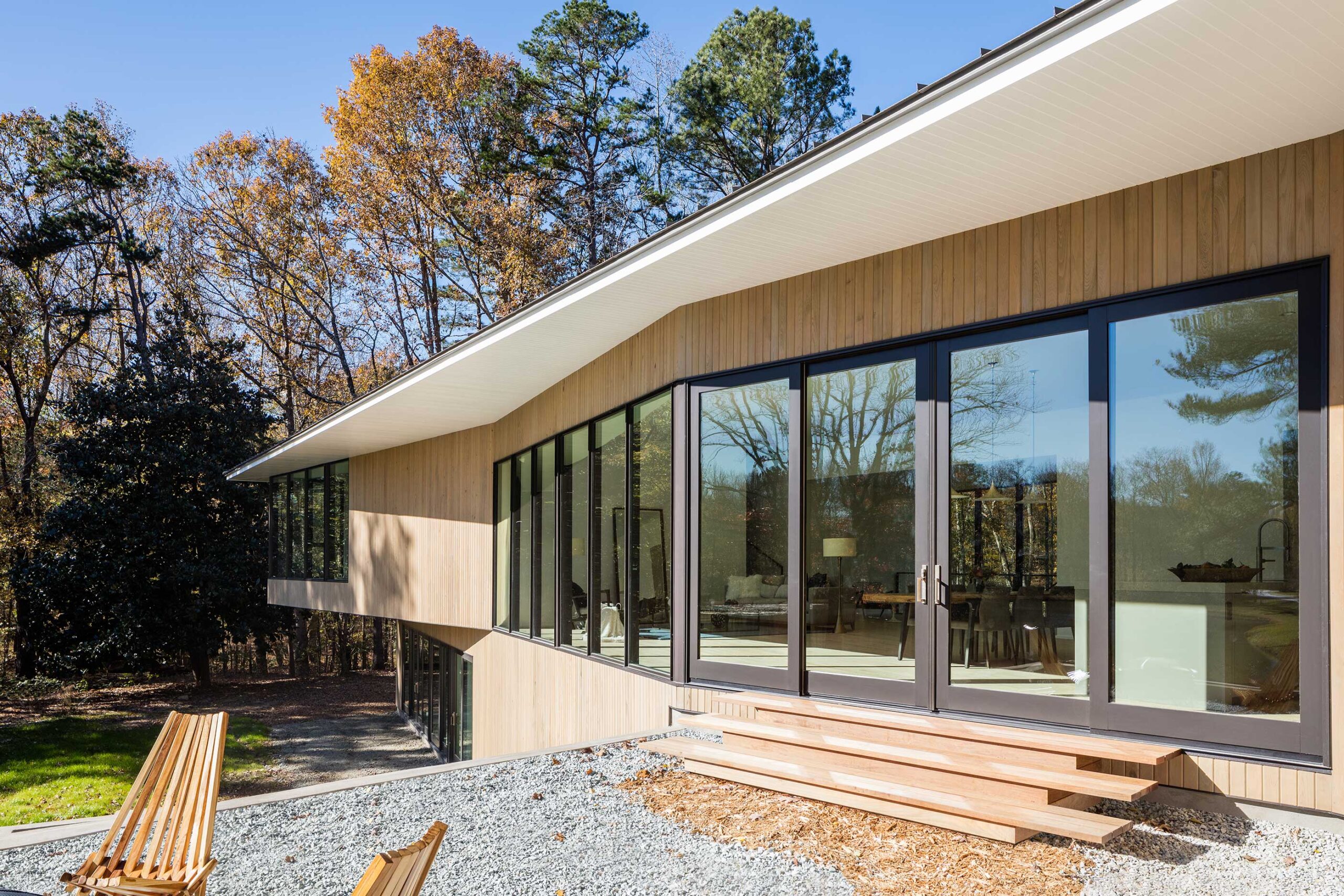 Modern house with wooden exterior, large glass windows, and a sloped roof, set in a wooded area. There are wooden benches and a gravel path in the foreground.