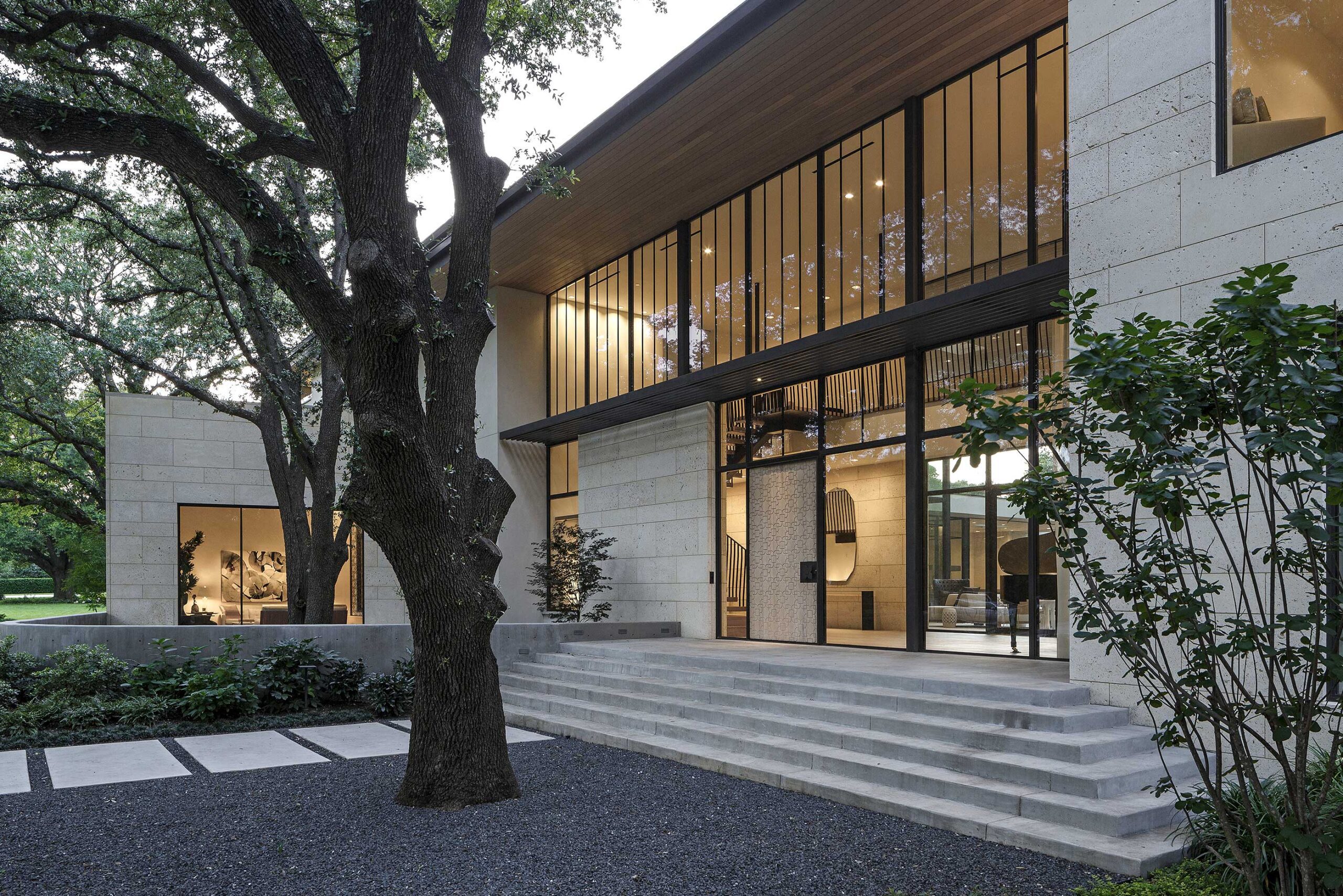 Modern house exterior with large windows, light stone walls, and a wide staircase. Surrounded by trees and greenery, the architectural design features clean lines and a minimalist aesthetic.