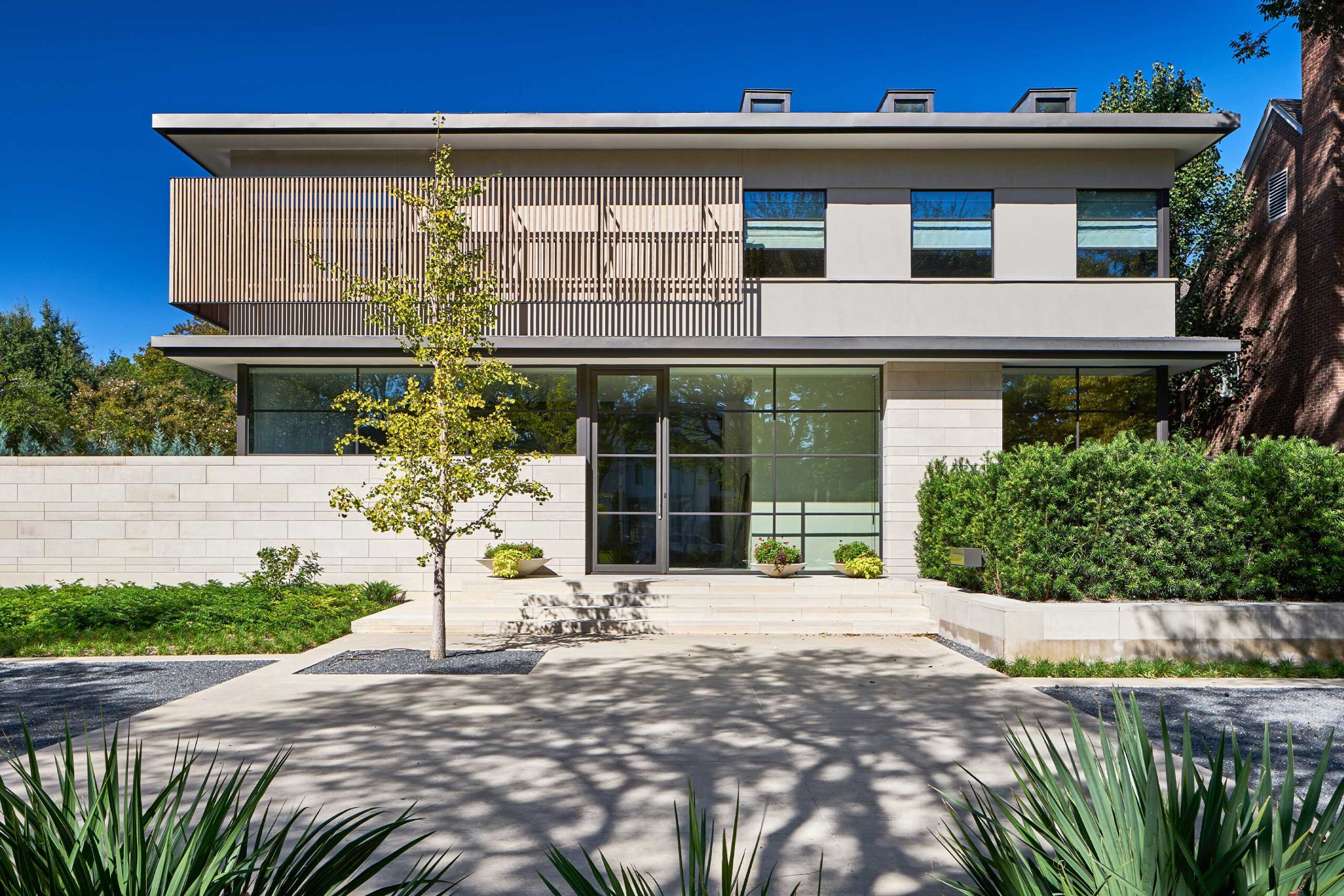 Modern two-story house with large windows, wooden panel accents, and a small tree in front. The driveway is bordered by plants and greenery.