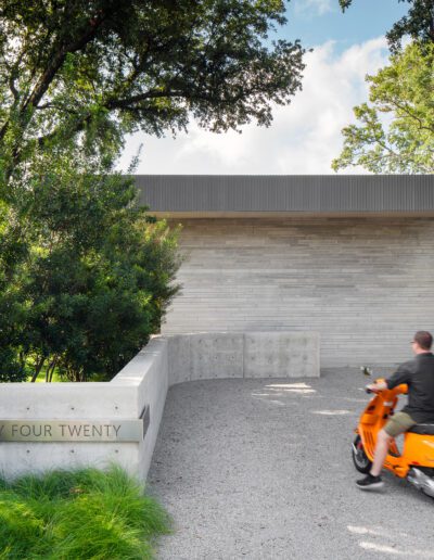 Person on an orange scooter approaches a modern house with light gray brick walls, surrounded by trees and greenery. A sign reads "forty four twenty.
