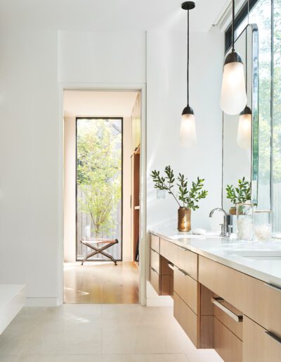 Modern bathroom with wood cabinetry, pendant lights, and a white countertop. A vase with green branches sits by the sink. A door leads to a small outdoor area with a bench and greenery.