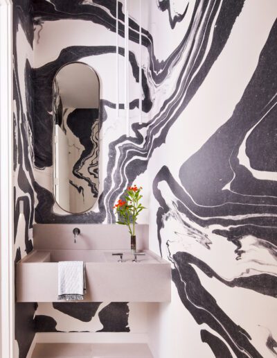 Bathroom with abstract black and white swirl-patterned walls, an oval mirror, floating sink, small vase with red flowers, and a hanging towel.
