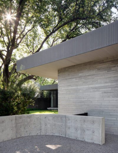 Modern building with a flat roof, textured concrete walls, and a curved concrete barrier in the foreground. Sunlight filters through trees in the background.