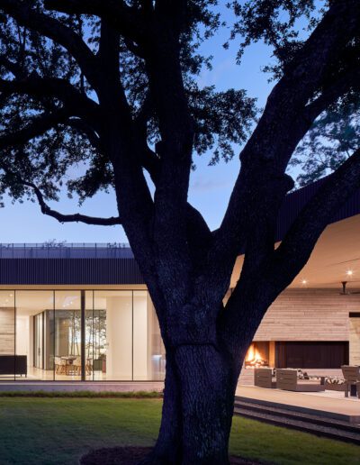 Modern house with large glass windows and a covered outdoor seating area, framed by a large tree in the foreground, during dusk.