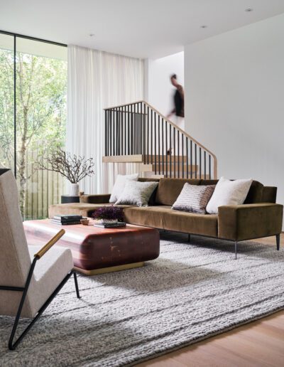 Modern living room with a brown sofa, wooden coffee table, and beige armchair. A person walks downstairs in the background. Large window with sheer white curtains and a view of greenery.