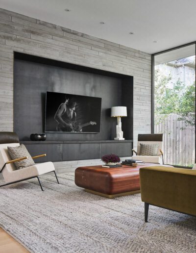 Modern living room with a large window, two beige armchairs, a green sofa, a wooden coffee table, and a wall-mounted TV on a gray brick wall. Rug covers light wood flooring.