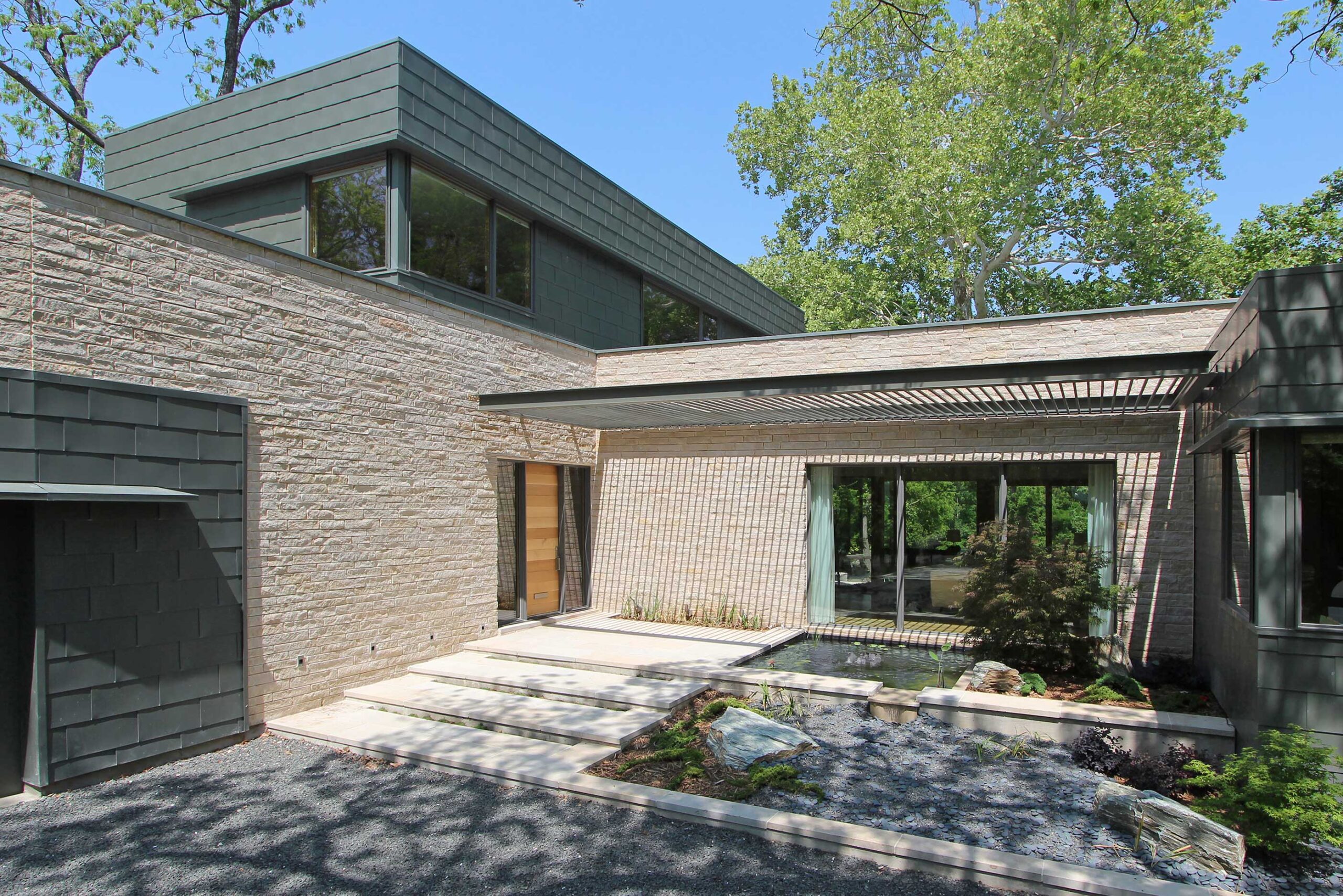 Modern house with gray stone walls and large windows, surrounded by trees. A shaded patio area and gravel pathway lead to the entrance.