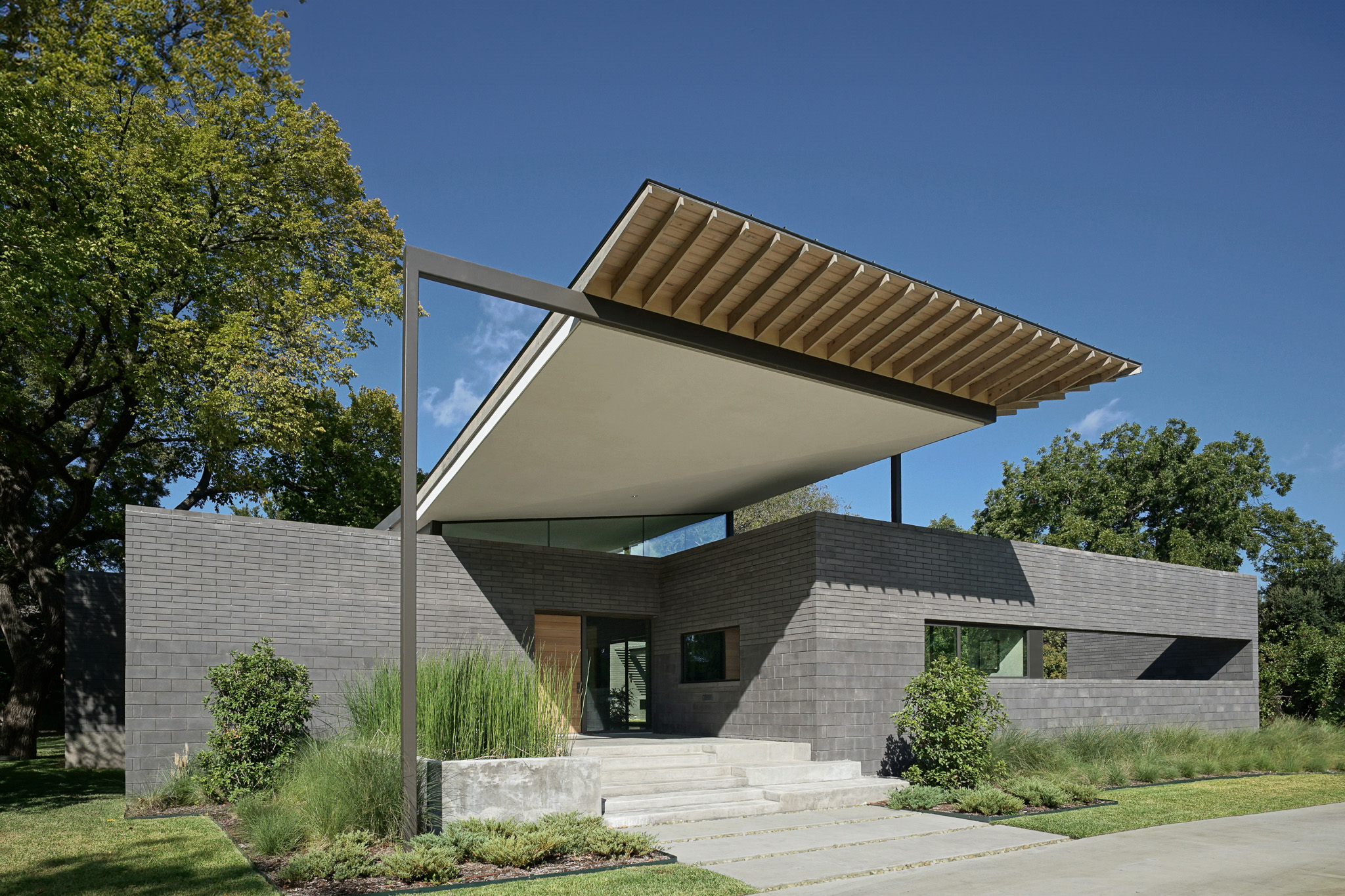 Modern house with geometric design and cantilevered roof, featuring a brick facade and large windows, surrounded by lush trees.