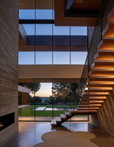 Modern interior with floating wooden staircase, large floor-to-ceiling windows, brick walls, built-in fireplace, and view of a lawn and trees at sunset.
