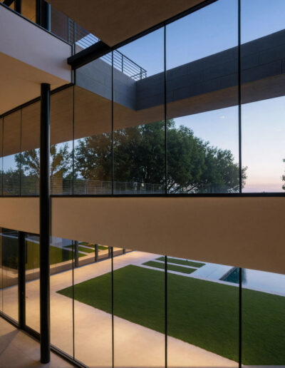 Modern building with large floor-to-ceiling glass windows overlooking a landscaped courtyard with grass, trees, and a swimming pool at dusk.