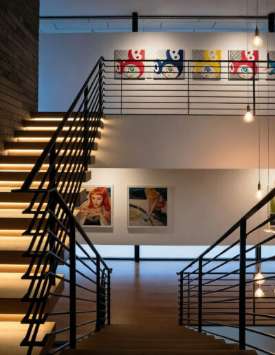 Modern staircase with illuminated steps, black railings, and contemporary artwork on the walls; hanging light bulbs add ambient lighting to the space.